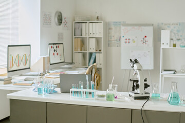 Well-lit science laboratory with computers, glassware, microscopes, and books neatly organized on tables and shelves surrounding the workspace