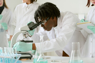 African American student examining samples under microscope while classmates taking notes in laboratory environment with lab equipment and glassware
