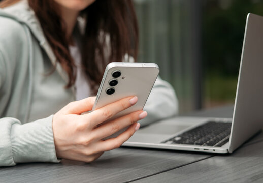 A person using a smartphone while sitting with a laptop on a wooden table outdoors, symbolizing multitasking and modern technology.