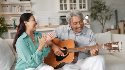 Happy senior man sitting at sofa and playing guitar or music instrument while his wife singing and...