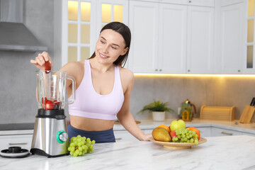 Woman in fitness clothes making delicious smoothie with blender at white marble table indoors