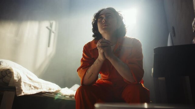 Young male inmate in orange uniform sitting on bed, praying deeply to God with hands clasped in sunlit prison cell with cross on wall in background