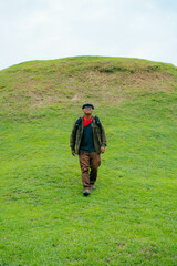 A male adventurer is enjoying the beauty of a small hill covered in green grass. The Asian male traveler is relaxing on the clean and beautiful green hills.