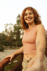 Smiling red-haired woman reaching forward on a wooden bridge in a natural park. Candid lifestyle...