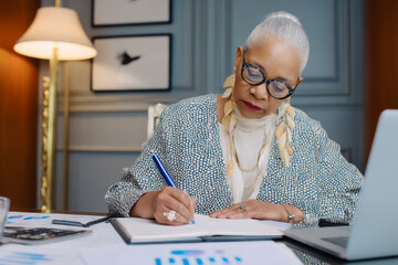 Elderly African-American woman reviewing quarterly earnings charts, making notes and preparing analysis in an elegant office