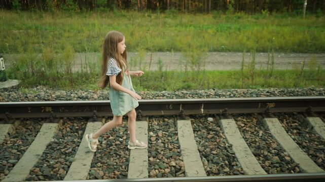 side view of young girl with long flowing hair walking confidently along rail track in countryside, wearing striped top and denim dress, surrounded by greenery and rocky railway bed in warm daylights