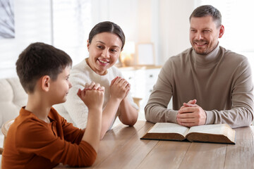 Family with Bible praying at table indoors