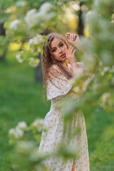 Beautiful Girl with long hair in a white dress poses near white blossoming apple trees