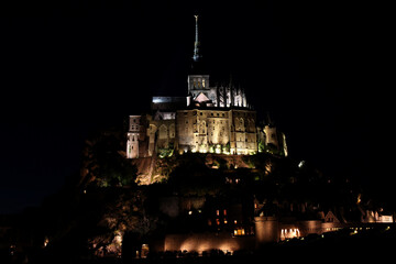 Night scene of Mont St Michel in France, with buildings lit by street lights and the tower lit by a search light, Normandy, northern France