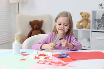 Happy little girl making handmade project with color paper and scissors at table indoors