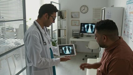 Side view of young confident radiologist in eyeglasses and lab coat communicating with patient while presenting him x-ray image of lungs and rib cage in clinics - Powered by Adobe