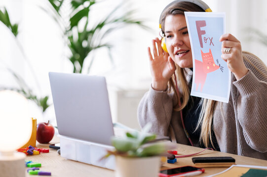 A teacher is leading a virtual class, showing a flashcard with a fox to help students learn the alphabet. She's using a laptop and headphones for the lesson.