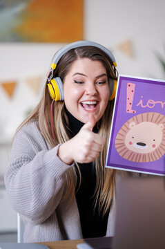 A teacher is giving a thumbs up while teaching the letter L with a lion flashcard. She's likely teaching online, hence the headphones.