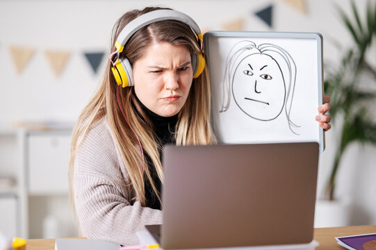 A woman with headphones is video conferencing, looking displeased. She's holding up a whiteboard with a drawing of a face that mirrors her expression, perhaps to illustrate her feelings.