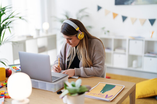 A woman with headphones is working at a desk. She's using a laptop and writing, possibly studying or working from home in a bright, organized space.