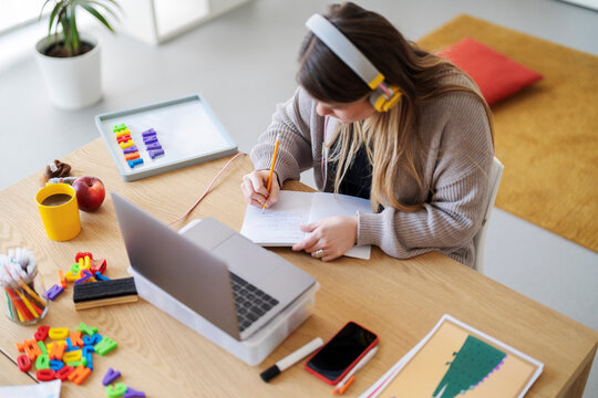 A teacher is planning lessons at her desk. She's using her laptop and writing in a notebook to prepare for her students. The colorful learning materials show her dedication..