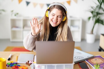 A teacher is waving during a virtual lesson. She's using a laptop and wearing headphones to connect with her students. She's teaching from home, surrounded by learning materials.