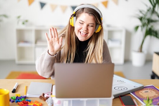 A teacher is waving during a virtual lesson. She's using a laptop and wearing headphones to connect with her students. She's teaching from home, surrounded by learning materials.