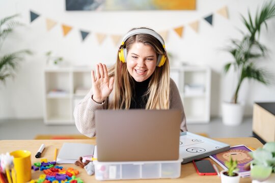 A teacher is waving during a virtual class. She's using a laptop and headset to connect with her students. She's teaching from home, surrounded by learning materials..