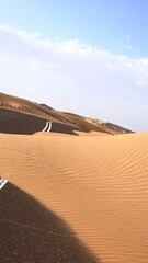 Sand dunes slowly reclaiming desert road under soft clouds, white road markings submerged by sands signify natural forces reclaiming manmade paths, inevitable nature resilience concept.