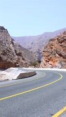 Road cuts clean line into curved desert hills, surrounded by compressed rocky walls stacked like books, glowing with layered mineral colors under midday sunlight. Canyon corridor concept.