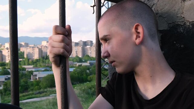 Portrait of a shaved-head teen boy in a prison cell, leaning his forehead against metal bars. He regrets his crime and reflects on his life. Juvenile detention, justice and redemption concept.