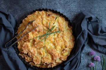 A potato gratin topped with cheese is placed on a table. A cloth is wrapped around the baking dish. Chives decorate the gratin. The picture is taken from above.