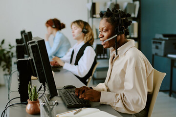 Woman wearing headset working in customer service call center with colleagues typing at computers in modern office environment