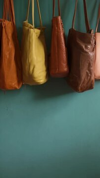 Collection of colorful leather tote bags hanging in a row against a teal textured wall on display.