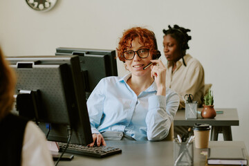 Portrait of woman smiling while working in customer service call center, wearing headset and sitting at desk with computer and coffee cup in foreground
