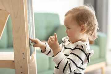 little kid girl drawing on black chalkboard in playroom, scratching doodles with colorful chalks,...