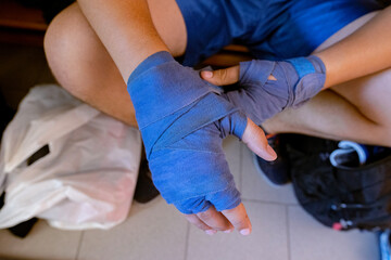 Athlete wrapping blue hand wraps around the left hand while seated, preparing for boxing or martial arts training. The focused action highlights the importance of wrist and hand protection.