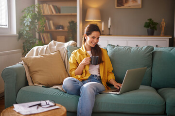 A woman dressed in a yellow shirt sits comfortably on a couch, sipping from a mug while focused on her laptop. The room has natural light and a relaxed atmosphere.
