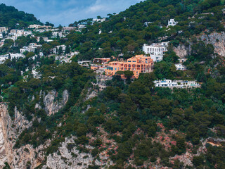 View of a vibrant orange villa on a steep hillside in Capri, Italy, surrounded by greenery, whitewashed buildings, and rugged Mediterranean cliffs. © Aerial Film Studio