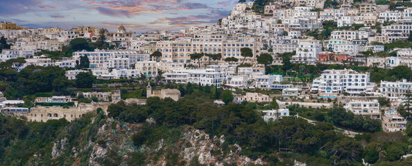 Densely packed whitewashed buildings with a prominent dome, lush greenery, and rocky cliffs on Capri Island, Italy, during sunset. © Aerial Film Studio