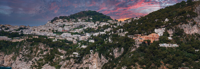 Capri Island's hillside with whitewashed buildings, lush greenery, and cliffs descending to the sea. A sunset sky adds pink and purple hues. © Aerial Film Studio