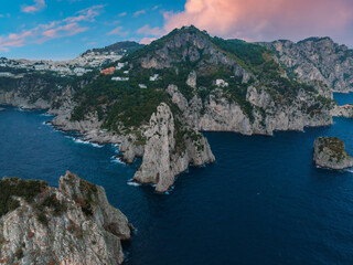 Fototapeta premium Capri Island's coastline features rocky cliffs, jagged sea stacks, lush greenery, and white hillside buildings under a pink and blue sky.