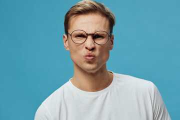 Obraz premium Close-up of a young man with short brown hair wearing glasses and a white t-shirt, making a funny face with puckered lips against a plain teal background, studio portrait. People lifestyle concept