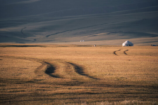 Tire tracks weave through golden grasslands in the Song Kol region, with yurts dotting the horizon under a sunset sky.