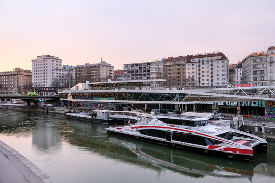 Urban landscape of the Donaukanal ports in Leopoldstadt, with the Twin City Liner terminal and moored catamarans along the waterfront, framed by cycling paths and riverside bars.