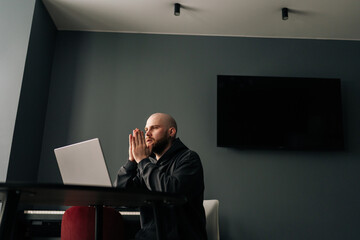 Remote view of focused male software developer with hands clasped, contemplating solution while working on laptop computer in a modern home office room, demonstrating deep concentration.