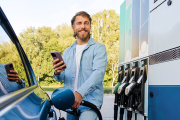 Handsome man at gas station filling up his car with one hand and checking smartphone with the other, smiling at camera