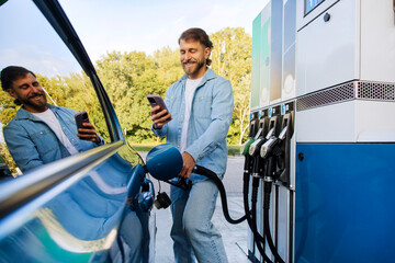 Happy handsome man refueling his car and messaging on smartphone, filling up automobile with bio...