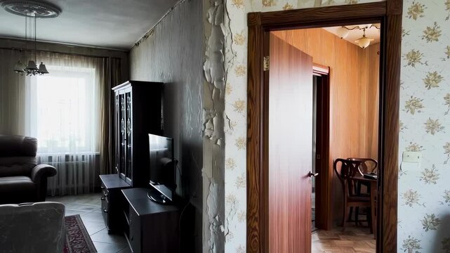Static scene inside a worn apartment. The living space has dark furnishings, a window with sheer curtains, and a noticeably peeling wall.