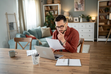 Focused freelancer reading documents and working on laptop at home