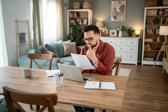 Focused freelancer reading documents and working on laptop at home