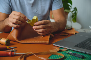 Craftsman gluing lining to brown leather bag in his workshop, surrounded by tools and laptop