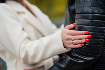 A couple embracing outdoors, a woman with red nails and a silver ring resting her hand on a man in a black leather jacket, suggesting romance and affection