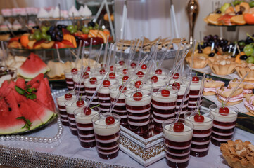 Festive dessert table display showcasing a wide assortment of sweet treats including layered strawberry and white mousse in glasses topped with cherries, watermelon slices, and pastries