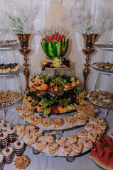 Elaborate dessert buffet display at a wedding reception featuring tiered stands with fruit arrangements, pastries, and cakes, adorned with gold accents and white drapery